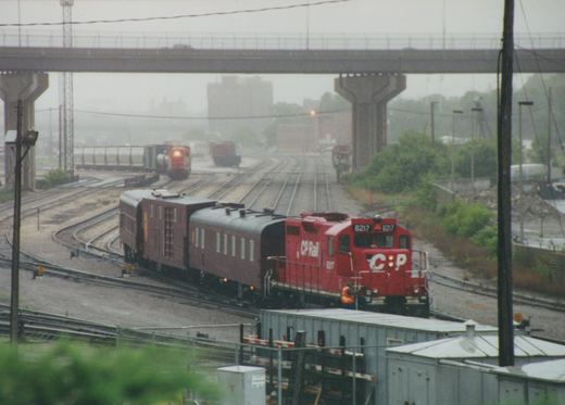A red Canadian Pacific locomotive numbered 8217 is pulling two maroon passenger cars on wet tracks under a concrete highway overpass on a rainy day.