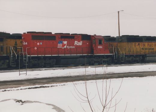 A red diesel locomotive with 'Rail System' lettering and the number 6084 sits on a track in a snowy yard, with a yellow and black Union Pacific locomotive beside it.