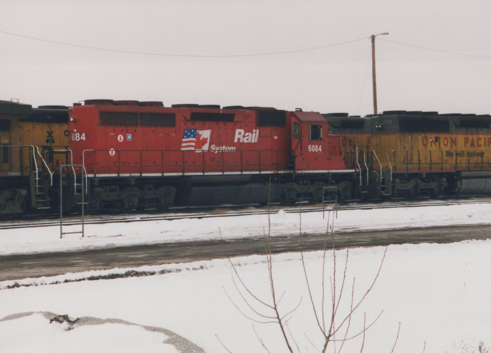 A red diesel locomotive with 'Rail System' lettering and the number 6084 sits on a track in a snowy yard, with a yellow and black Union Pacific locomotive beside it.