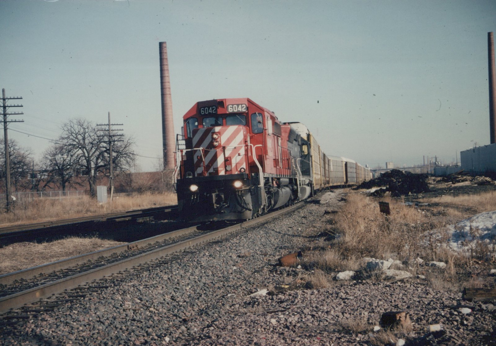 A red and white Canadian Pacific diesel locomotive numbered 6042 pulls a line of freight cars along a railroad track in a winter industrial landscape.