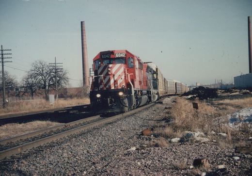 A red and white Canadian Pacific diesel locomotive numbered 6042 pulls a line of freight cars along a railroad track in a winter industrial landscape.