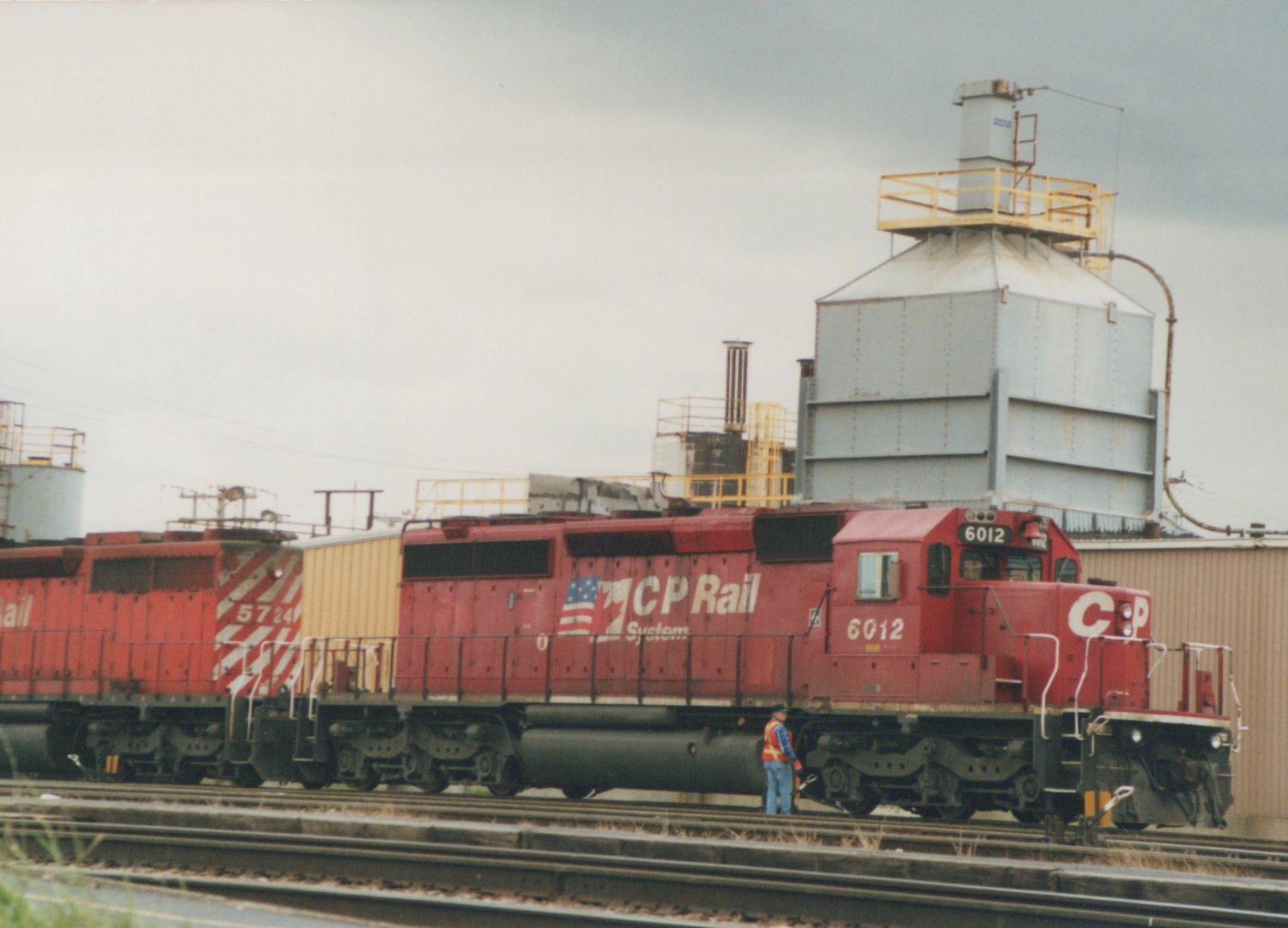 Two red diesel locomotives with CP Rail System lettering, numbers 6012 and 5724, are parked on tracks in a yard with industrial buildings in the background, under a cloudy sky.