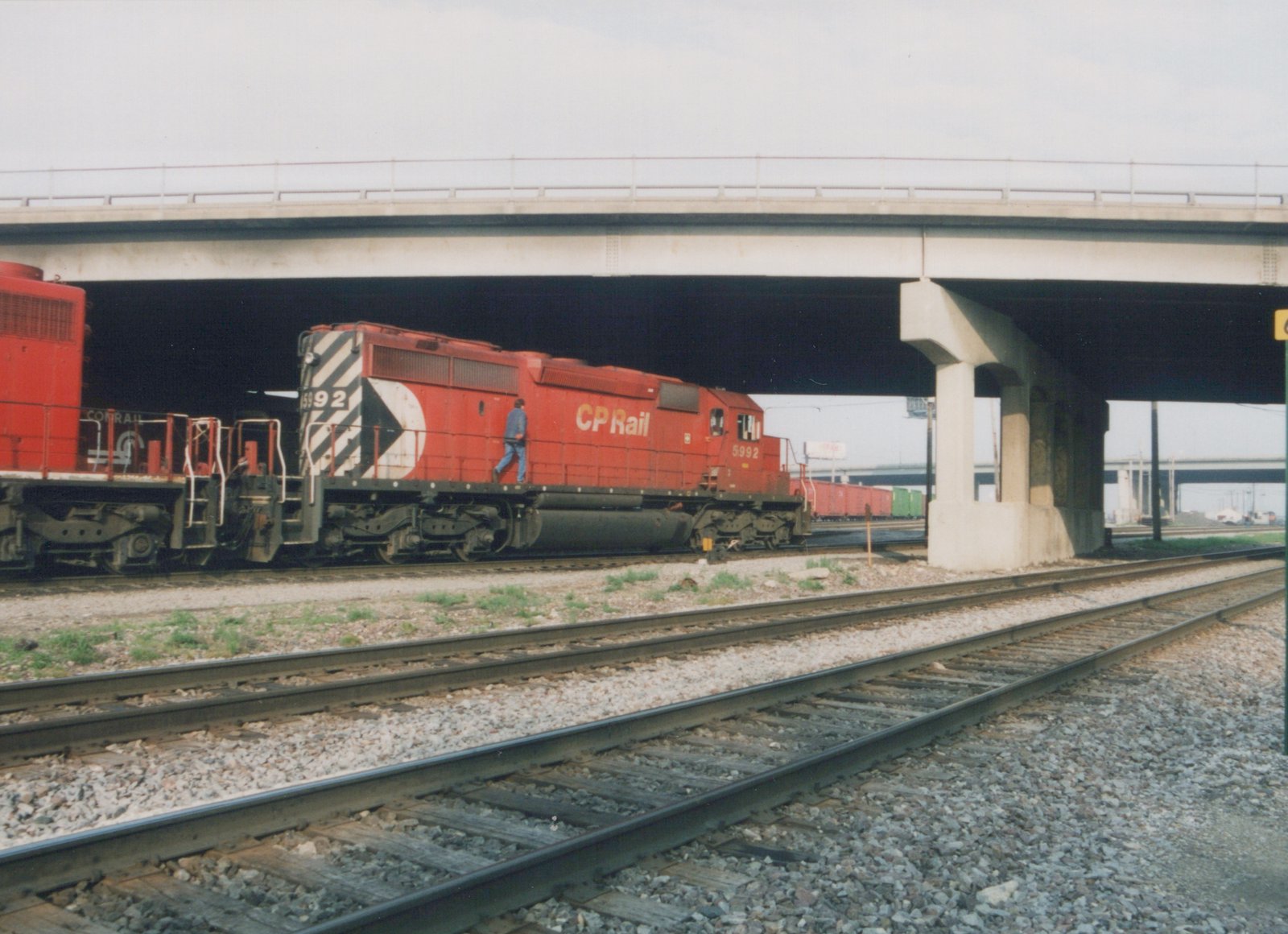 A side view of a red CP Rail diesel locomotive, number 5992, under an overpass with a Conrail unit behind it, on a set of railroad tracks in Franklin Park, Illinois.