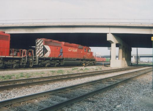 A side view of a red CP Rail diesel locomotive, number 5992, under an overpass with a Conrail unit behind it, on a set of railroad tracks in Franklin Park, Illinois.