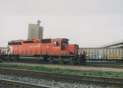 A red Canadian Pacific GP38-2 locomotive, number 5992, stands on the tracks with a gray freight car covered in graffiti behind it, under an overcast sky.