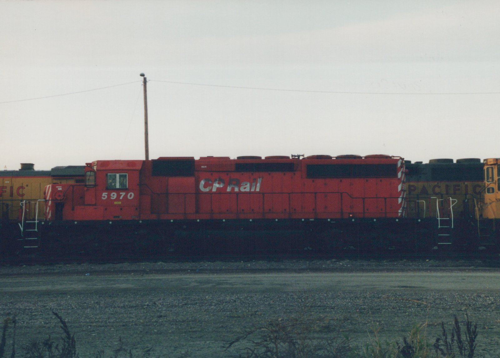 Side view of a red CP Rail diesel locomotive number 5970 in a gravel yard, with a yellow locomotive partially visible behind it, under an overcast sky.