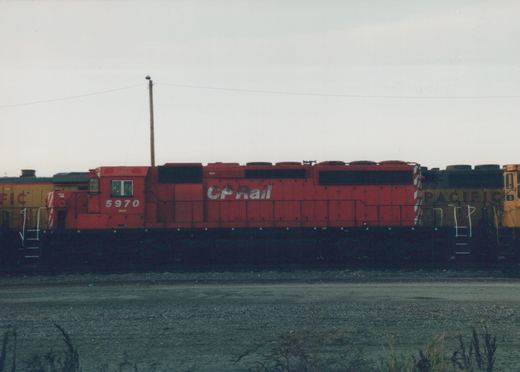 Side view of a red CP Rail diesel locomotive number 5970 in a gravel yard, with a yellow locomotive partially visible behind it, under an overcast sky.