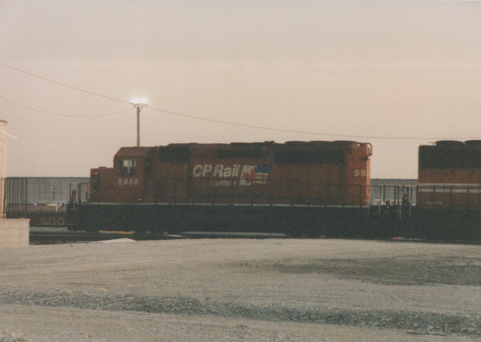 A red and black Canadian Pacific diesel locomotive numbered 5958 sits stationary in a rail yard with a gravel foreground and a light pole in the background.