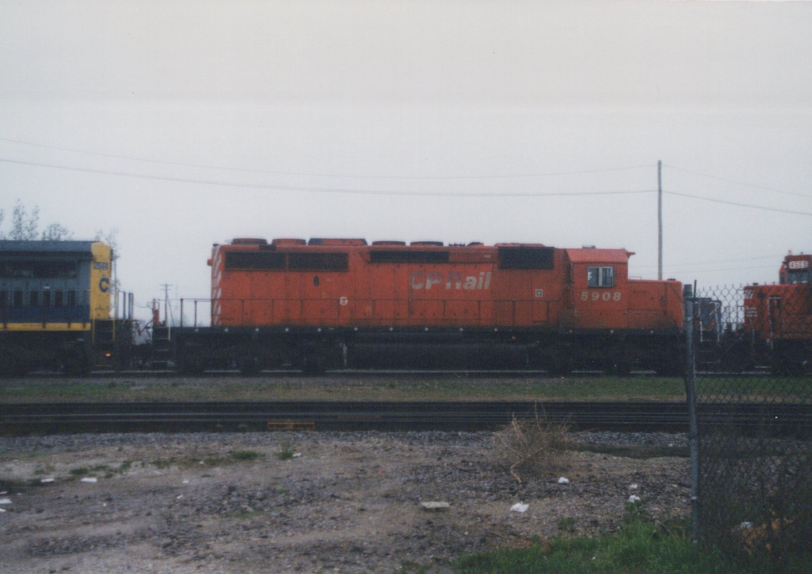 An orange CP Rail locomotive numbered 5908 is parked on a track in a gravel yard under an overcast sky, with a blue and yellow passenger car and another orange locomotive visible in the background.