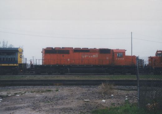 An orange CP Rail locomotive numbered 5908 is parked on a track in a gravel yard under an overcast sky, with a blue and yellow passenger car and another orange locomotive visible in the background.