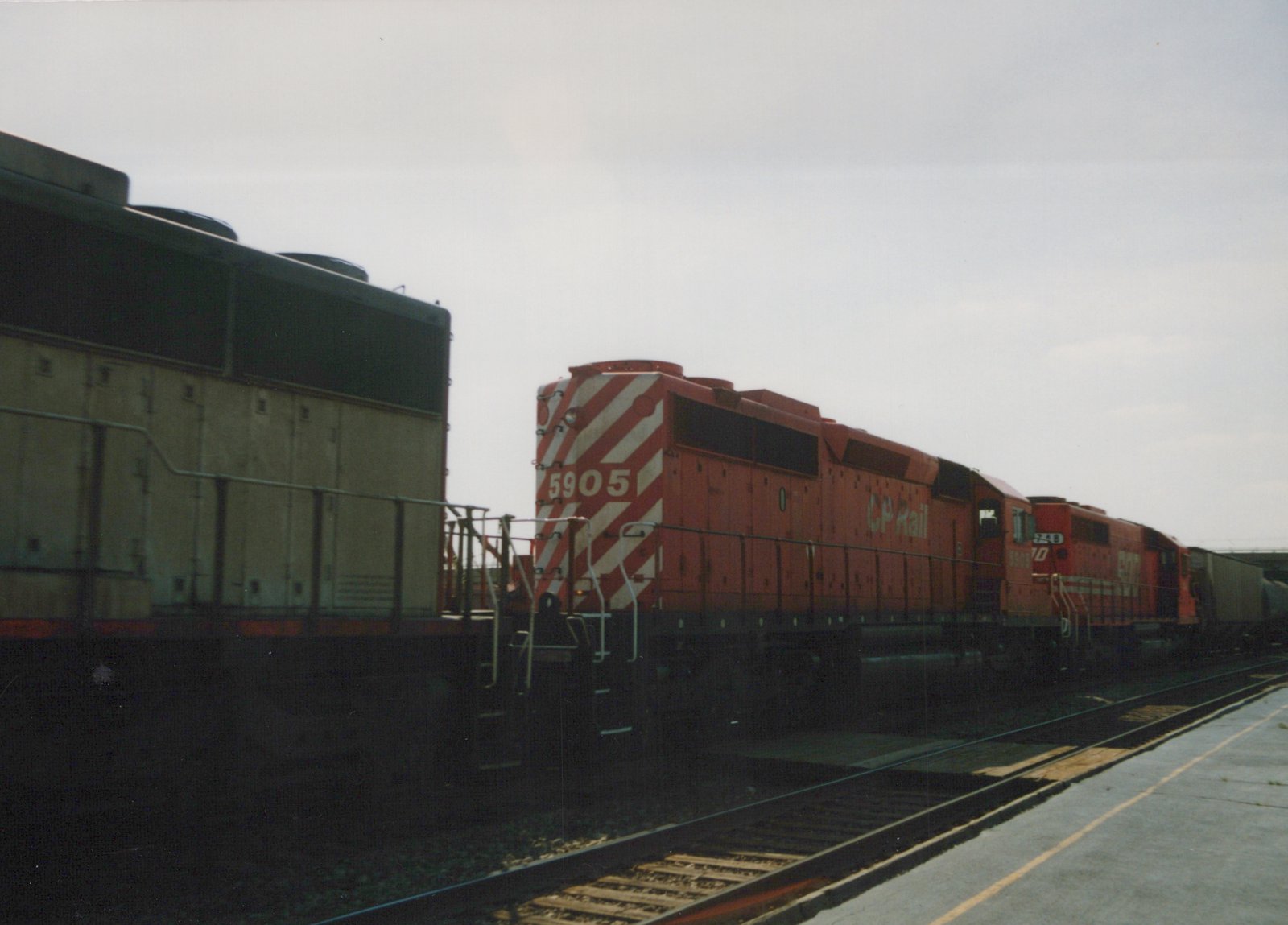 Two red CP Rail diesel locomotives, numbered 5905 and 5908, are parked on tracks beside a station platform under an overcast sky.