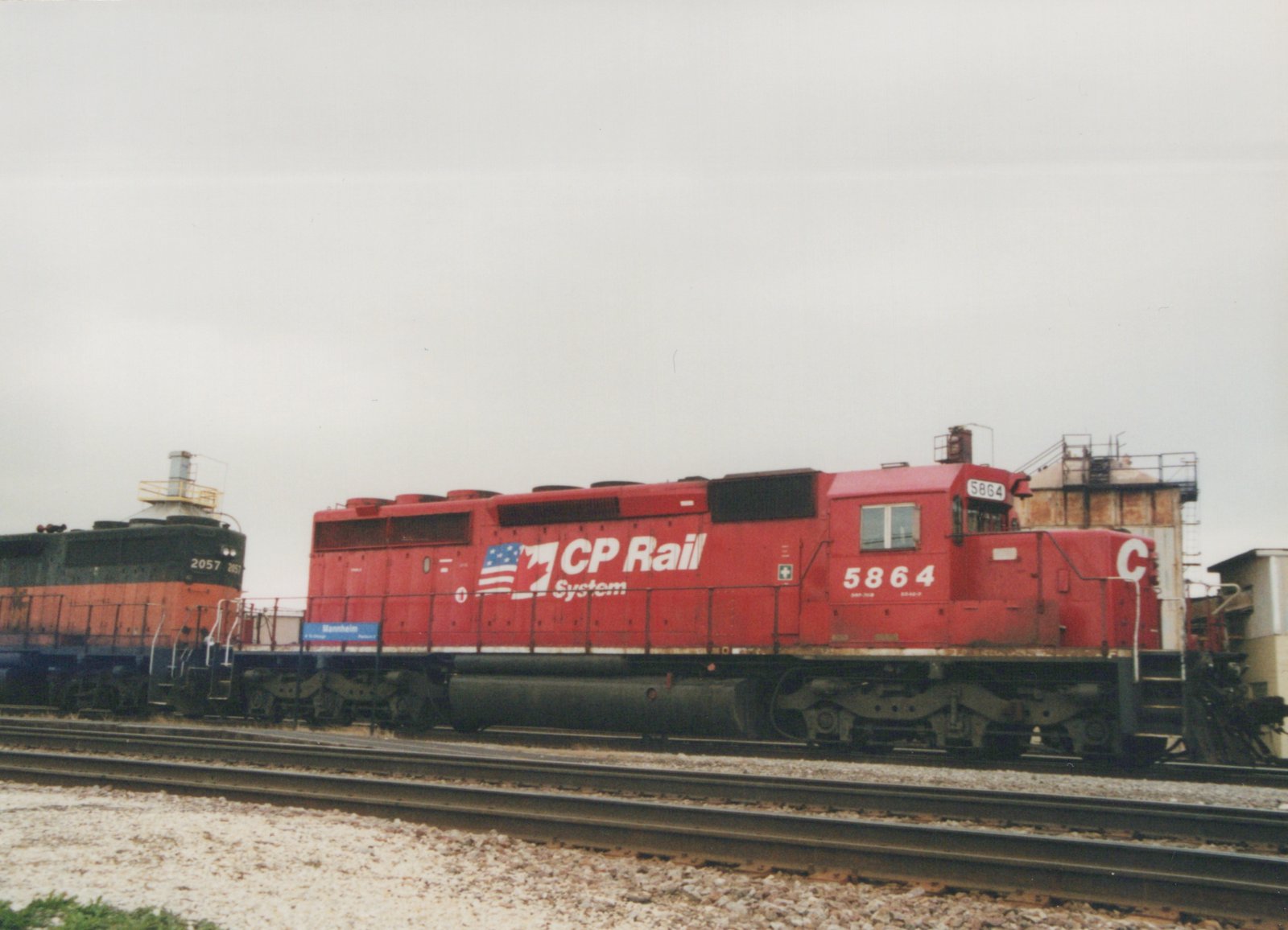 A red CP Rail locomotive numbered 5864 is parked on railroad tracks next to a dark green and orange Santa Fe locomotive numbered 2057, with a blue station sign reading 'Mannheim' in the background under an overcast sky.