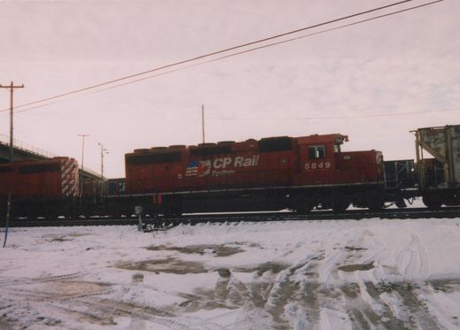 A red CP Rail System diesel locomotive, numbered 5849, is shown on railroad tracks beside a snow-covered field, with a second locomotive and freight cars behind it.