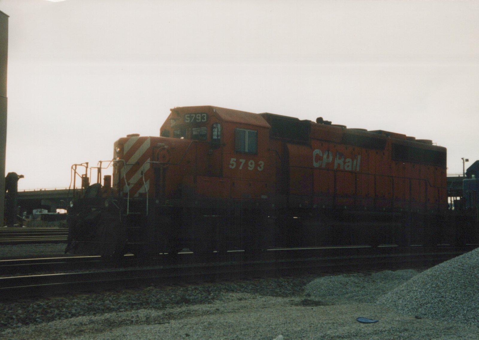 A side view of an orange CP Rail locomotive numbered 5793 on railroad tracks in a gravel yard under a cloudy sky.