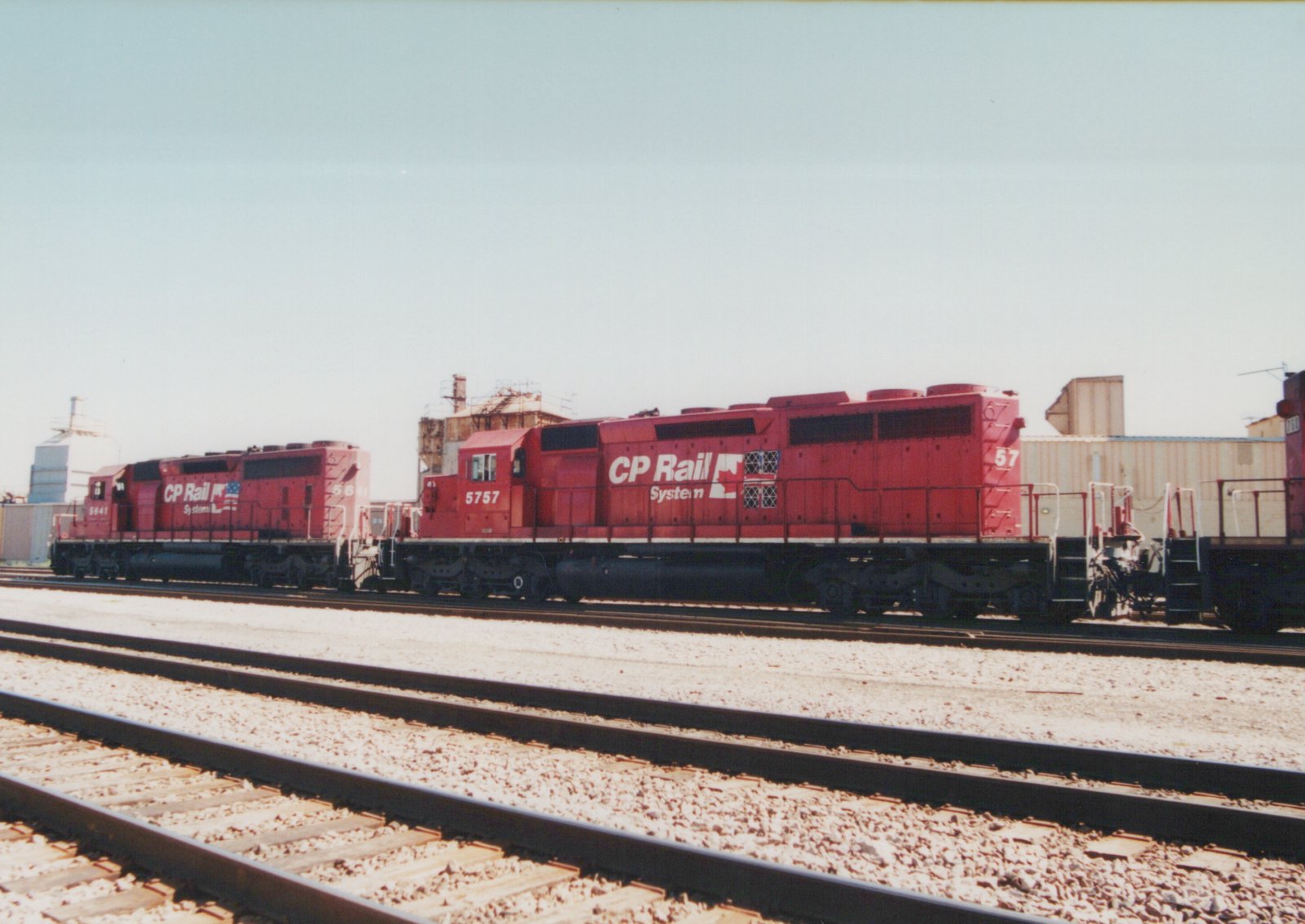 Two red CP Rail System diesel locomotives are shown parked on parallel tracks in a rail yard under a clear sky.