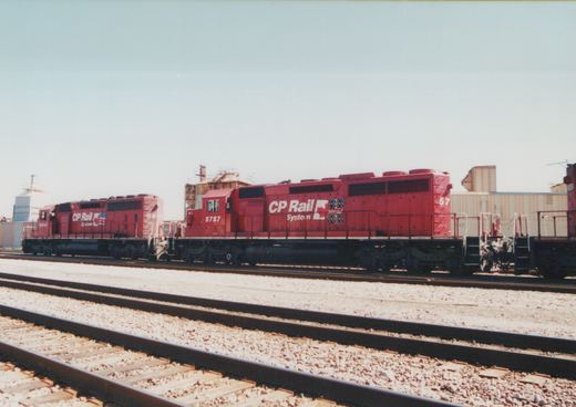 Two red CP Rail System diesel locomotives are shown parked on parallel tracks in a rail yard under a clear sky.