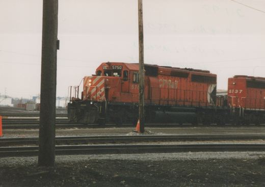 Two red CP Rail diesel locomotives, numbered 5750 and 5737, are positioned on parallel tracks in a rail yard, with utility poles and a traffic cone in the foreground.
