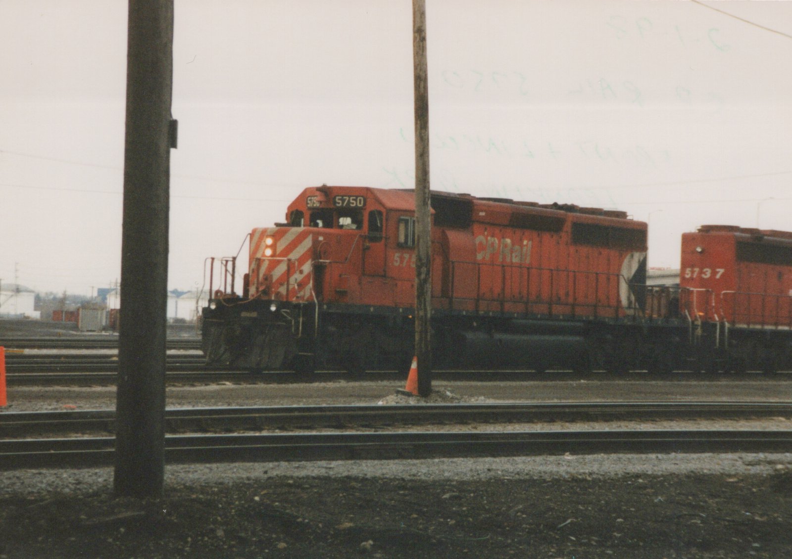 Two red CP Rail diesel locomotives, numbered 5750 and 5737, are positioned on parallel tracks in a rail yard, with utility poles and a traffic cone in the foreground.