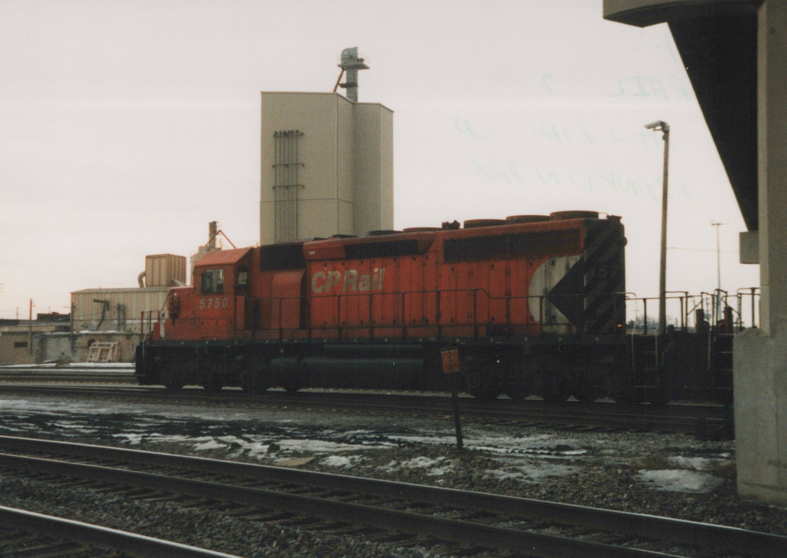 A red Canadian Pacific locomotive numbered 5750 is stationary on a railroad track in a snowy yard, with industrial buildings in the background.