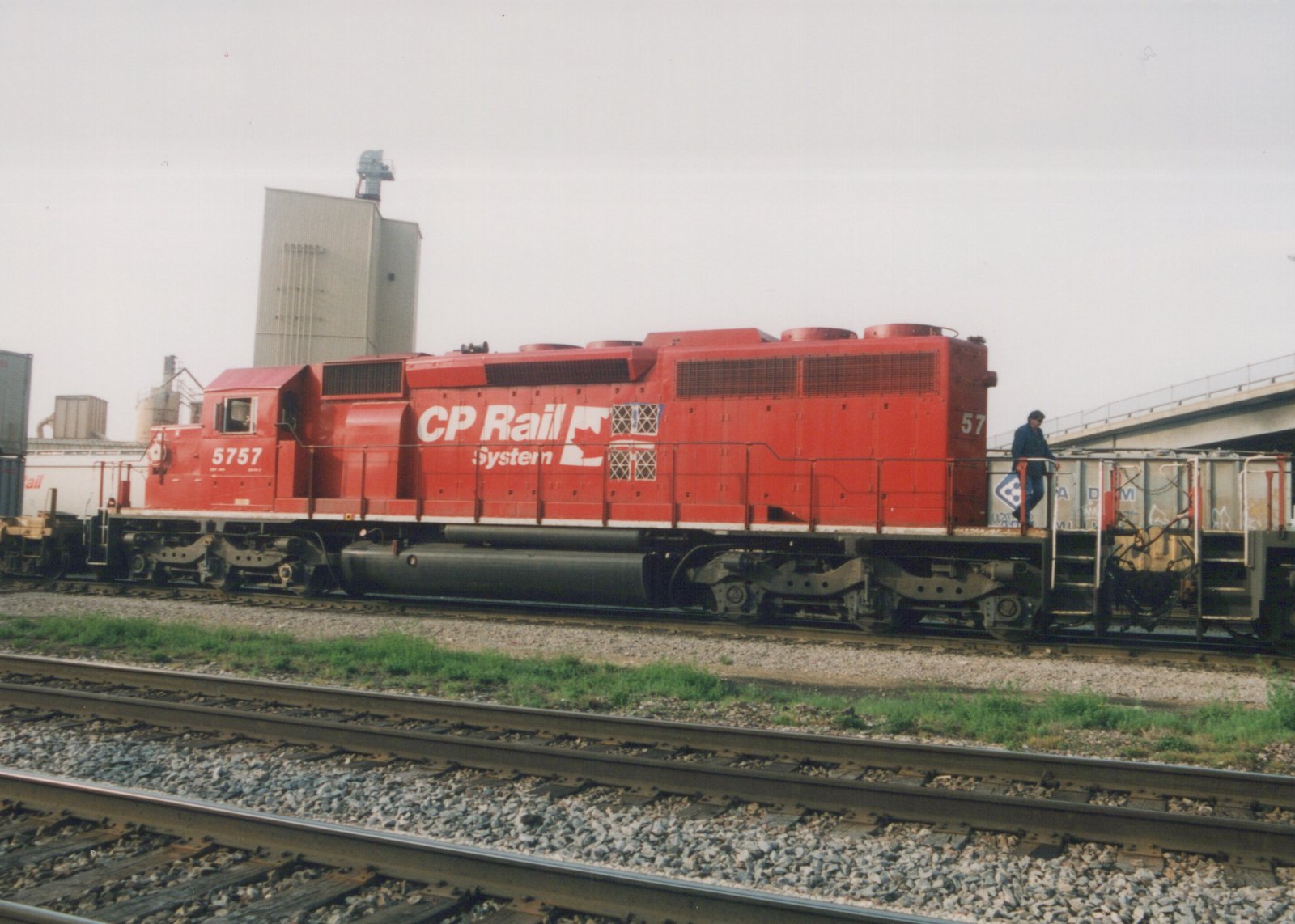 A side view of a bright red diesel locomotive with 'CP Rail System' and the number 5757 on its side, standing on a set of tracks next to another freight car, with a person on its rear platform, under an overcast sky.