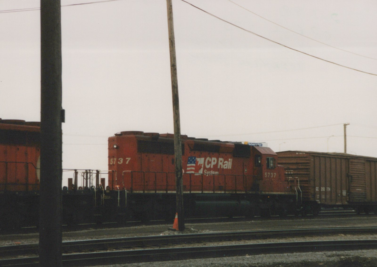A red CP Rail System diesel locomotive, number 5737, is shown on a railroad main line, pulling a brown freight car, with utility poles and an overcast sky in the background.