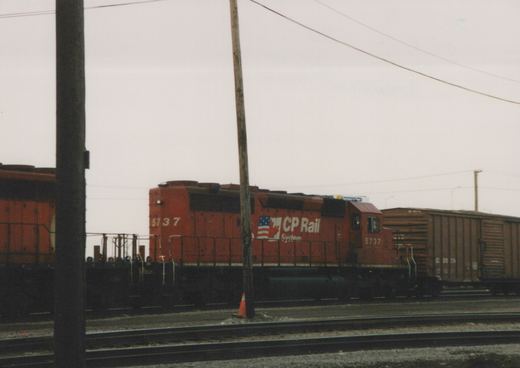 A red CP Rail System diesel locomotive, number 5737, is shown on a railroad main line, pulling a brown freight car, with utility poles and an overcast sky in the background.
