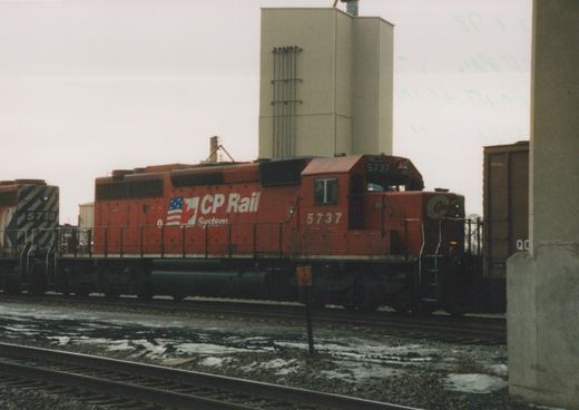A red CP Rail System diesel locomotive, number 5737, stands on a track in a snowy yard, with another locomotive and a freight car visible in the background.