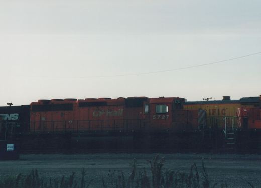 A side view of an orange CP Rail diesel locomotive numbered 5727, standing in a rail yard at dusk with a black Norfolk Southern freight car and a yellow locomotive in the background.