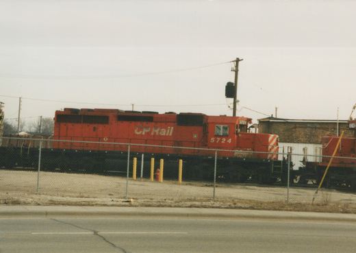 A red CP Rail diesel locomotive, number 5724, stands in a fenced yard on a cloudy day, with another red locomotive partially visible behind it.