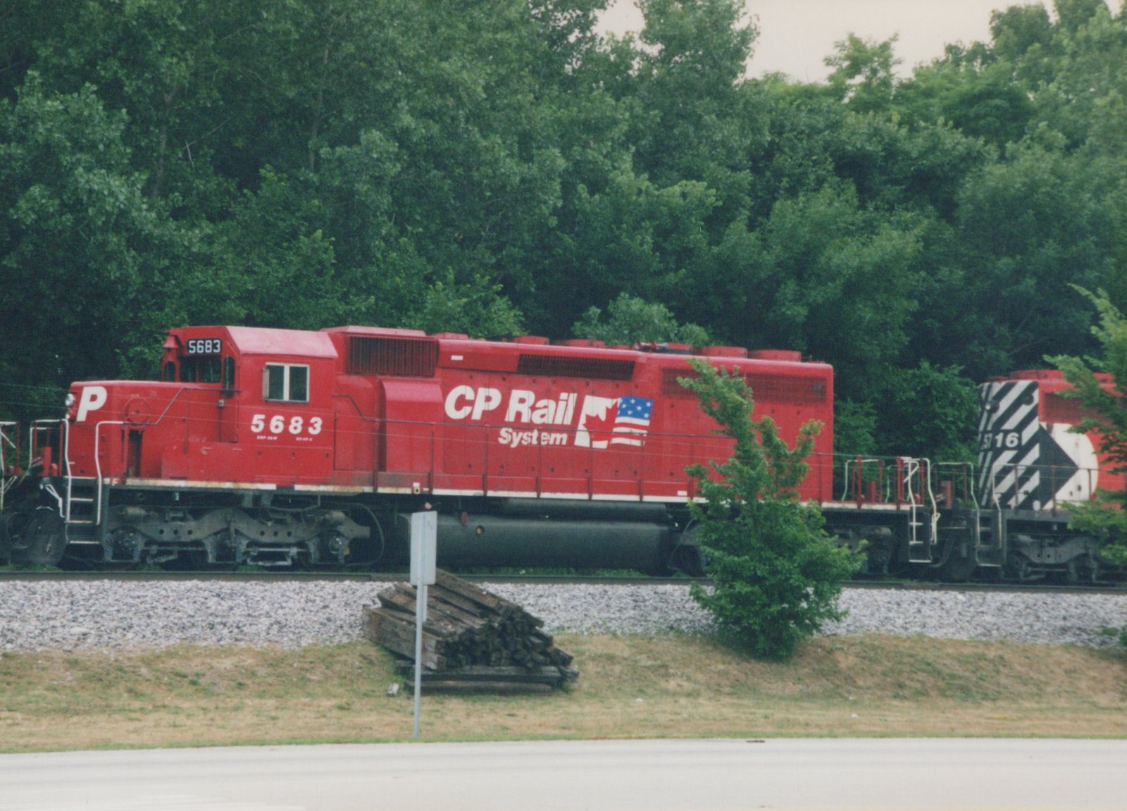 A bright red CP Rail System diesel locomotive numbered 5683 travels along tracks bordered by trees and grass, with a second locomotive, number 516, following behind.