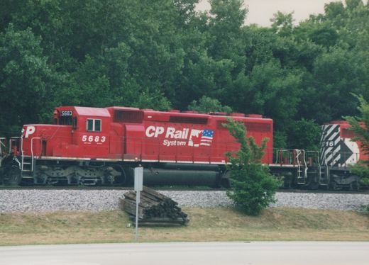 A bright red CP Rail System diesel locomotive numbered 5683 travels along tracks bordered by trees and grass, with a second locomotive, number 516, following behind.