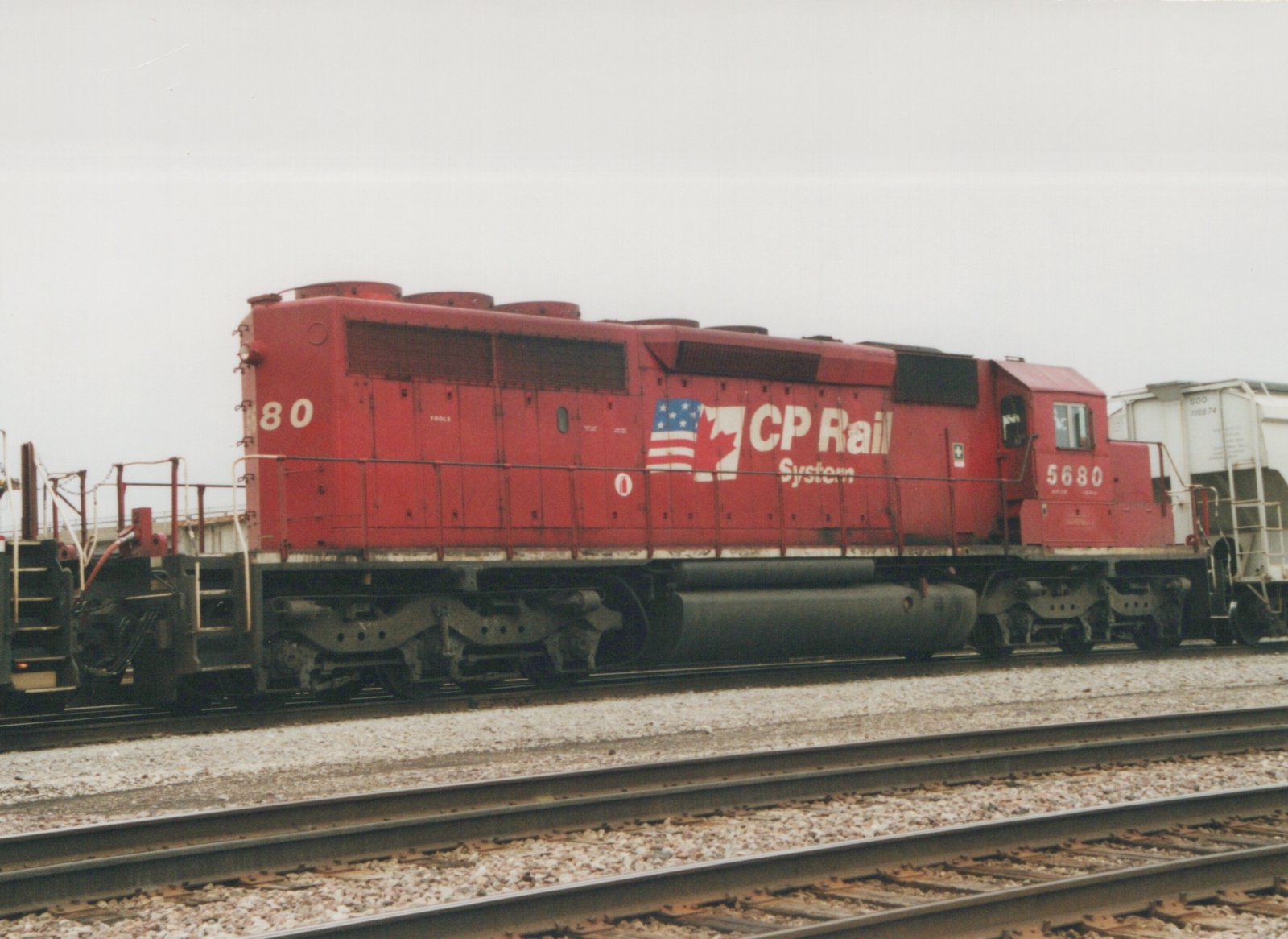 A red Canadian Pacific SD40-2 diesel locomotive, numbered 5680, is parked on a set of tracks in a railyard. The side of the locomotive features the CP Rail System logo with American and Canadian flags. The background includes a white freight car.