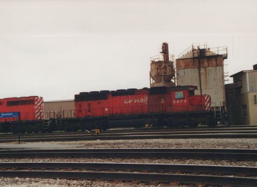 A red Canadian Pacific GP9 diesel locomotive, numbered 5674, stands on tracks near a station sign for Mannheim Platform 2, with industrial structures in the background.