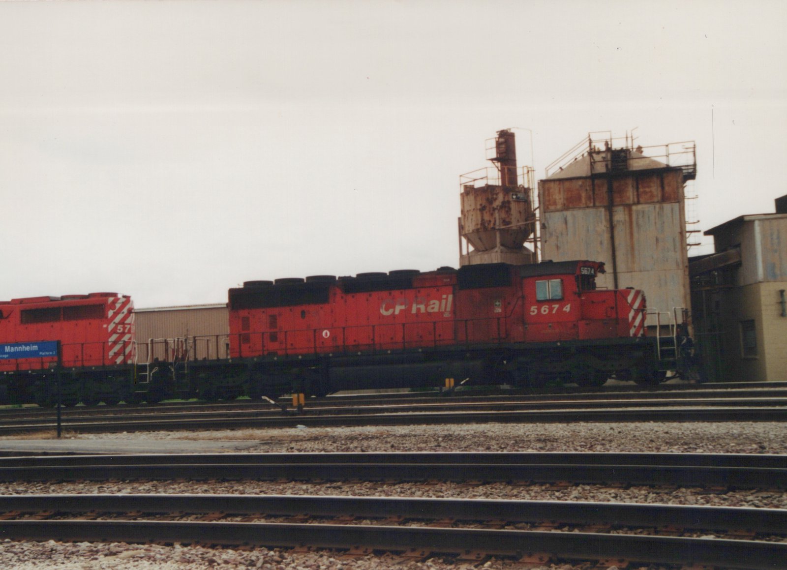 A red Canadian Pacific GP9 diesel locomotive, numbered 5674, stands on tracks near a station sign for Mannheim Platform 2, with industrial structures in the background.