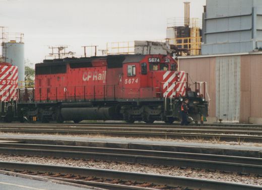 A red CP Rail System diesel locomotive numbered 5674 sits on tracks in a rail yard, with another locomotive numbered 5728 partially visible behind it. A person stands near the front of the locomotive.