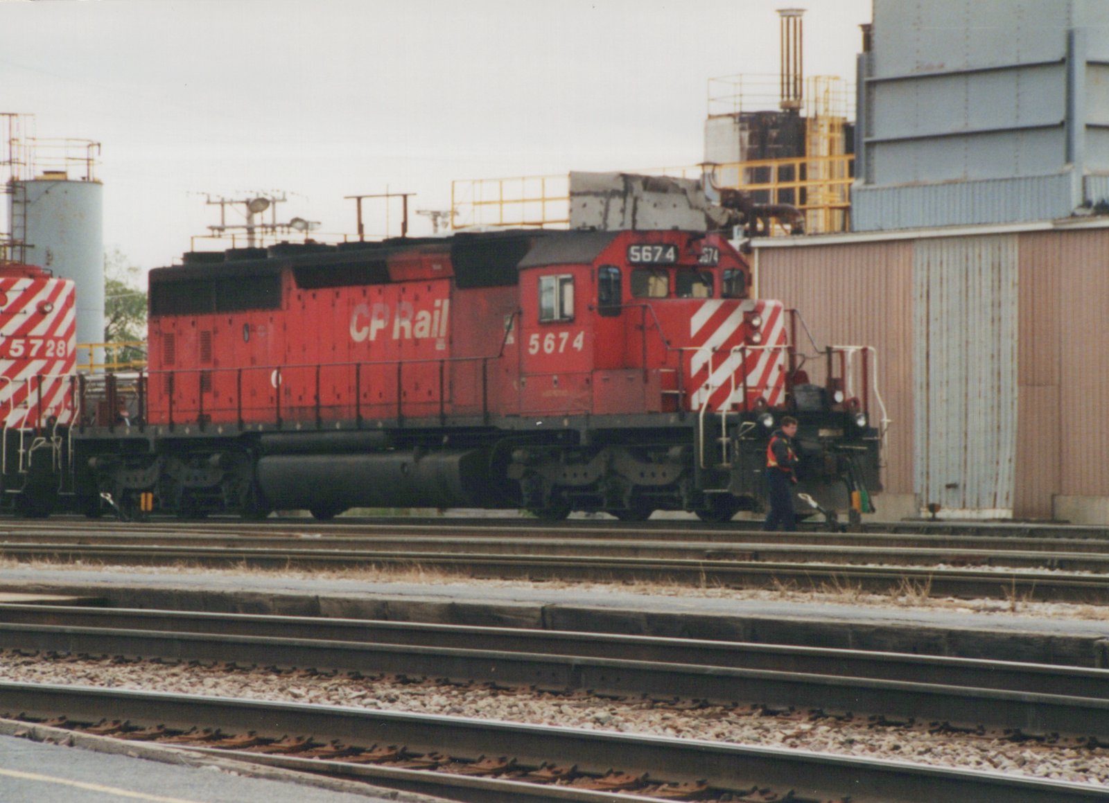 A red CP Rail System diesel locomotive numbered 5674 sits on tracks in a rail yard, with another locomotive numbered 5728 partially visible behind it. A person stands near the front of the locomotive.