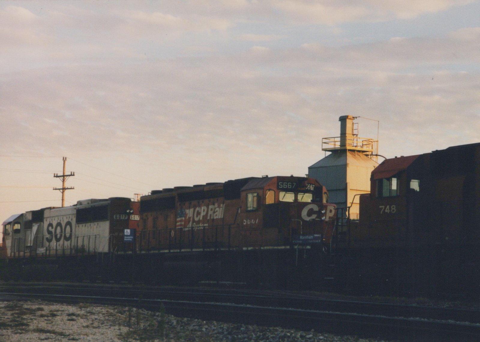 Three diesel locomotives from CP Rail System and SOO Line are parked side-by-side on tracks at a yard in Franklin Park, Illinois, with a grain silo and a 'Mannheim' station sign visible, under an overcast dusk sky.
