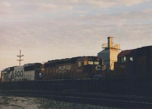 Three diesel locomotives from CP Rail System and SOO Line are parked side-by-side on tracks at a yard in Franklin Park, Illinois, with a grain silo and a 'Mannheim' station sign visible, under an overcast dusk sky.