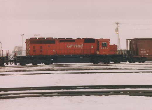 A red CP Rail diesel locomotive with the number 5674 is parked on tracks in a snowy yard, with a freight car visible to its right.