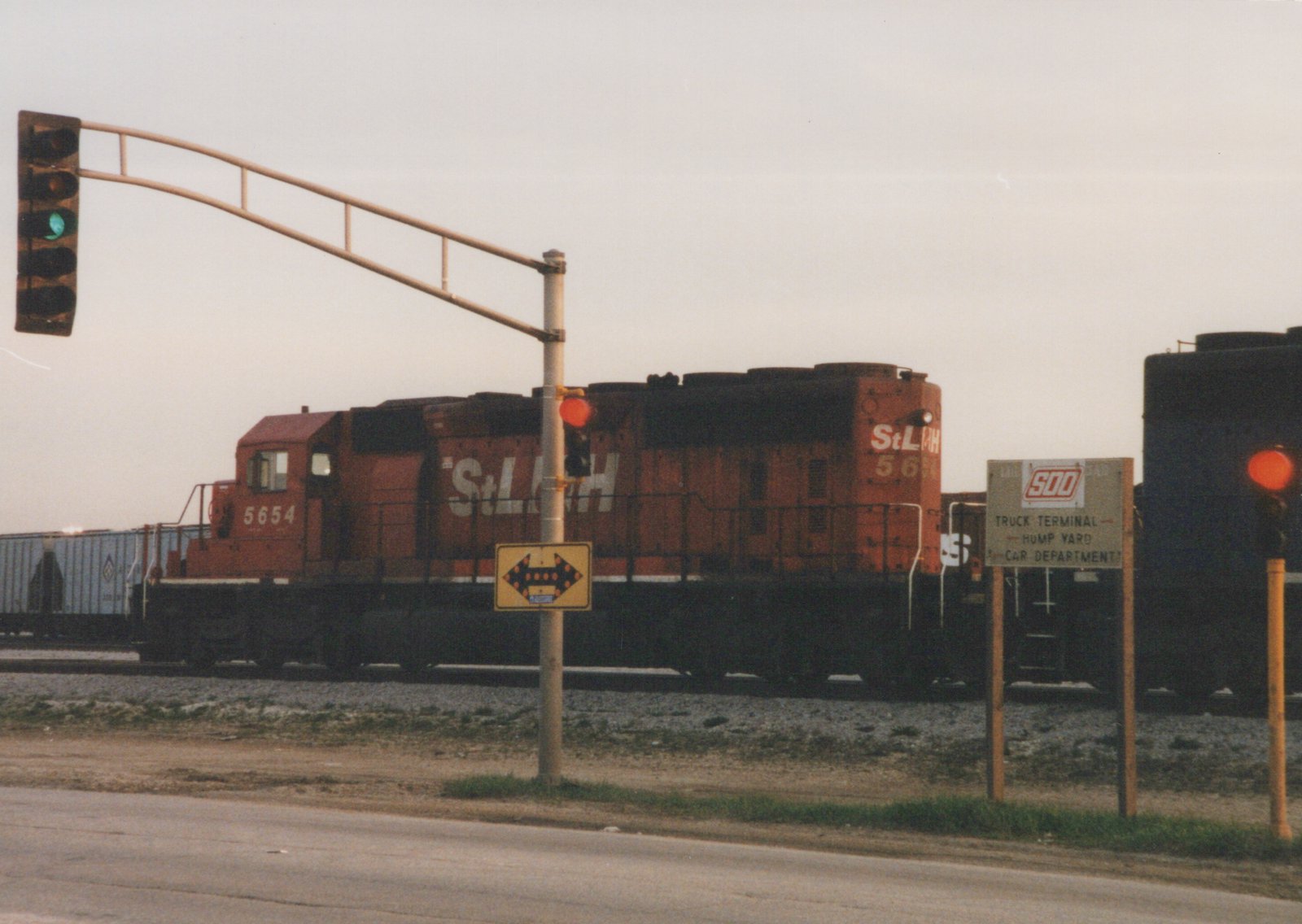 A red diesel locomotive numbered 5654 with StLH lettering is stopped at a street crossing with traffic lights, next to a sign for the SOO Truck Terminal.