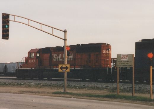 A red diesel locomotive numbered 5654 with StLH lettering is stopped at a street crossing with traffic lights, next to a sign for the SOO Truck Terminal.