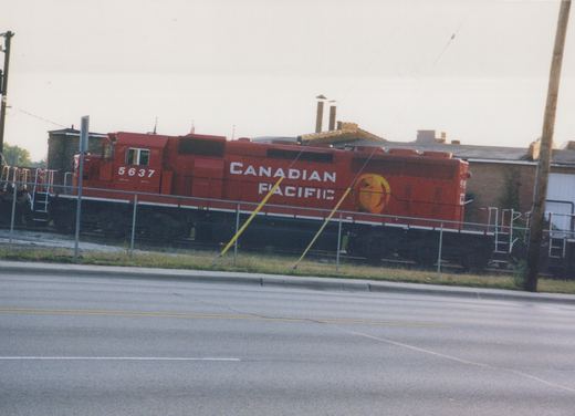 A red Canadian Pacific SD40-2 locomotive numbered 5637 sits on a track behind a chain-link fence, viewed from across a paved street.