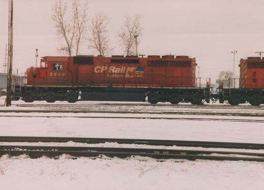 A side view of a red CP Rail System diesel locomotive, number 5549, on a snow-covered track in a rail yard, with a second red locomotive coupled behind it.