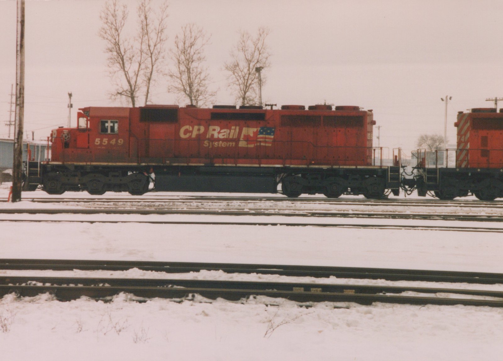 A side view of a red CP Rail System diesel locomotive, number 5549, on a snow-covered track in a rail yard, with a second red locomotive coupled behind it.