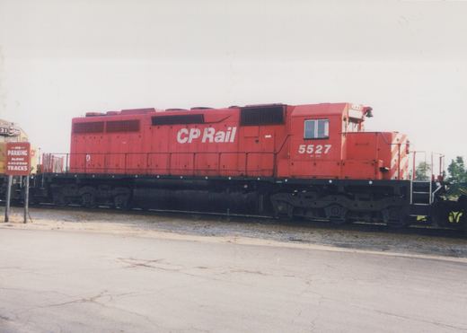 A bright red Canadian Pacific diesel locomotive is parked on a railroad track next to a cracked asphalt road, with a 'No Parking' sign visible.