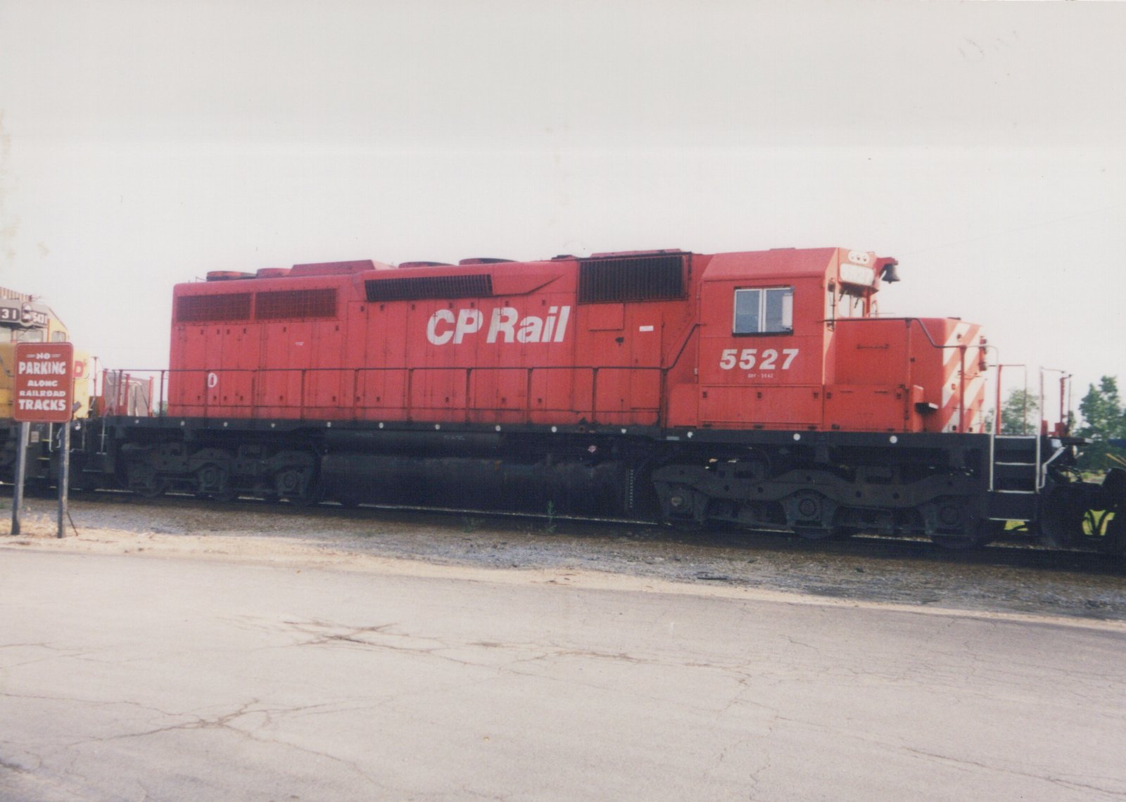 A bright red Canadian Pacific diesel locomotive is parked on a railroad track next to a cracked asphalt road, with a 'No Parking' sign visible.