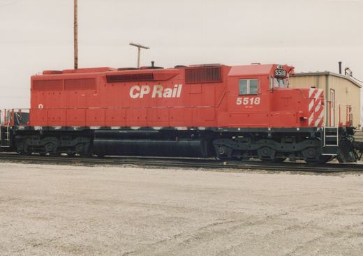 A red and white CP Rail diesel locomotive, number 5518, stands on tracks in a gravel yard, with its side fully visible and a building in the background.
