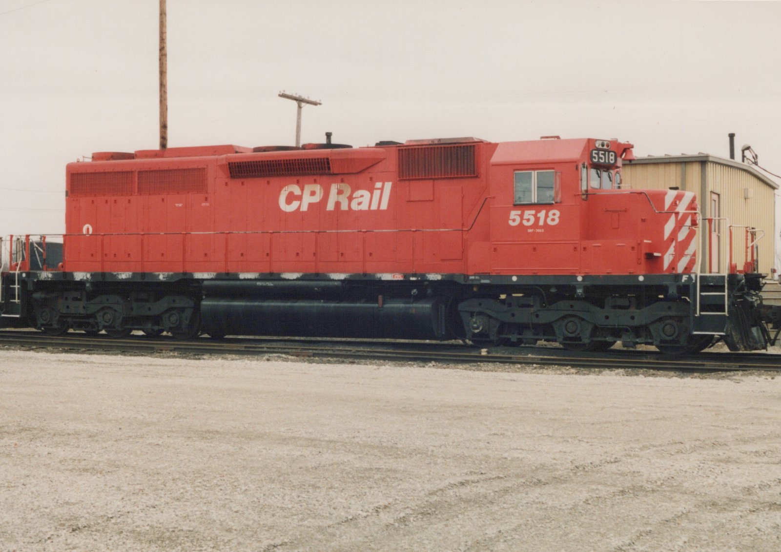 A red and white CP Rail diesel locomotive, number 5518, stands on tracks in a gravel yard, with its side fully visible and a building in the background.