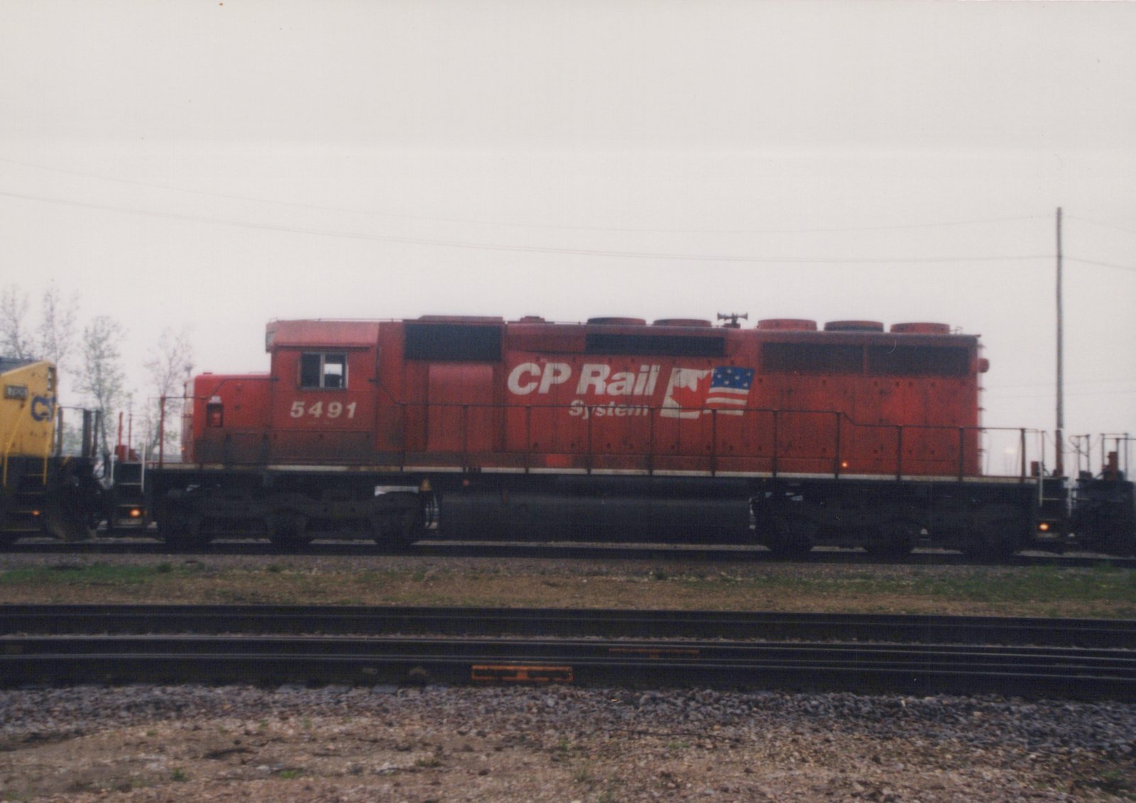 A red CP Rail System locomotive numbered 5491 is parked on railroad tracks, with a yellow locomotive partially visible to its left, under an overcast sky.