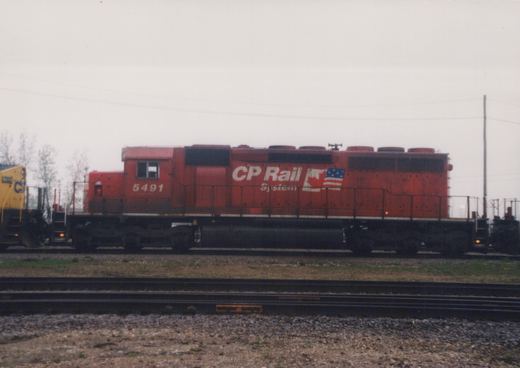 A red CP Rail System locomotive numbered 5491 is parked on railroad tracks, with a yellow locomotive partially visible to its left, under an overcast sky.